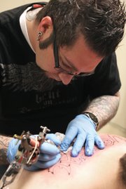 Jimmy Clark fills the sharp points of the lettering on Tucker Vestal’s back Thursday at Liquid Flesh Tattoo. Clark then switched instruments to fill the remainder of the tattoo with black and red coloring. Clark also uses rubber gloves, disinfectants and cleaners to guarantee a sterile working environment.